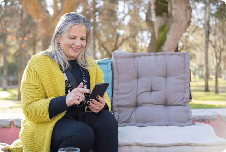 Frau mit grauem Haar und gelber Strickjacke sitzt im Freien und schaut lächelnd auf ihr Smartphone.